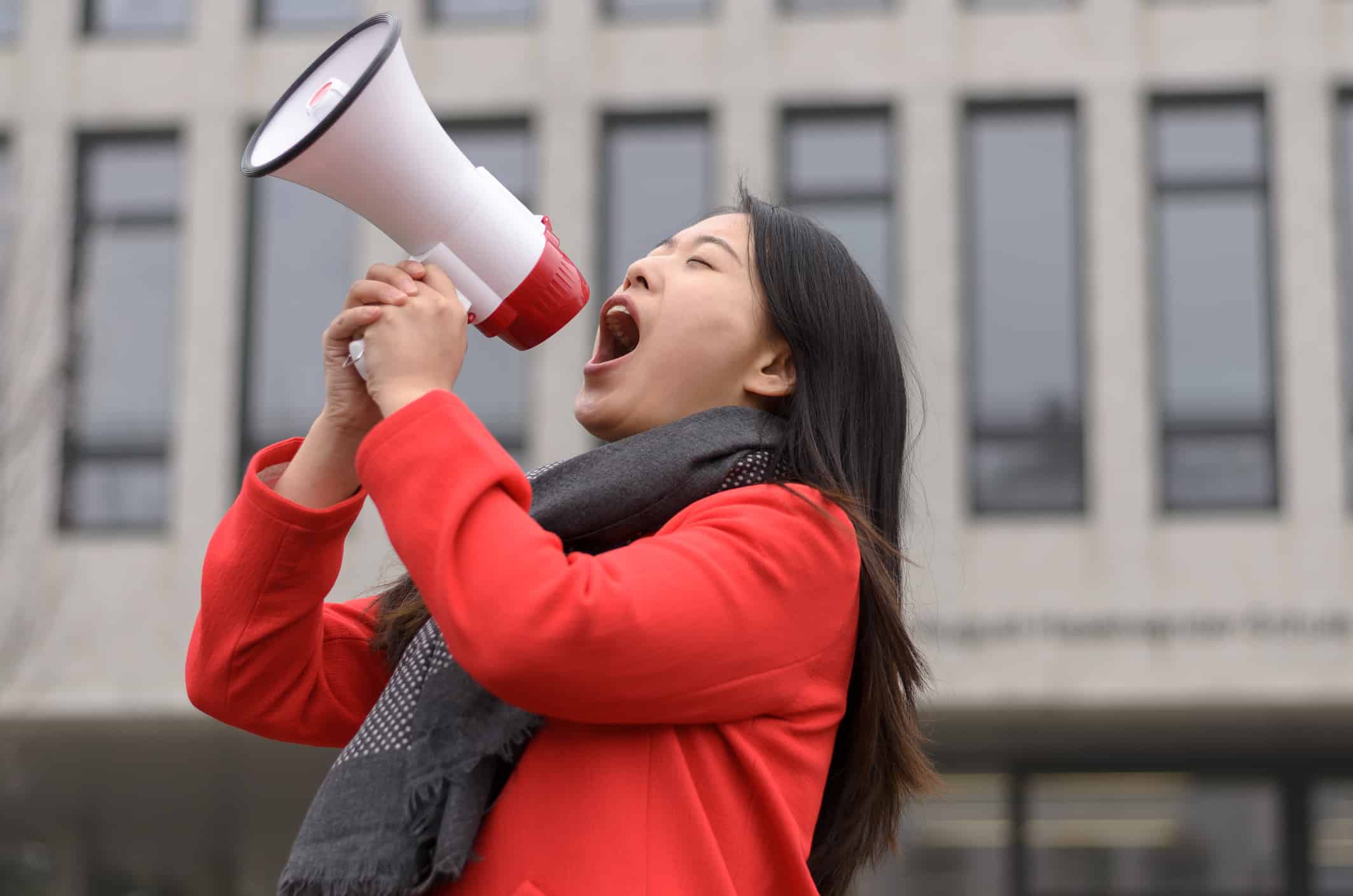 Asian woman in red coat shouting into megaphone.