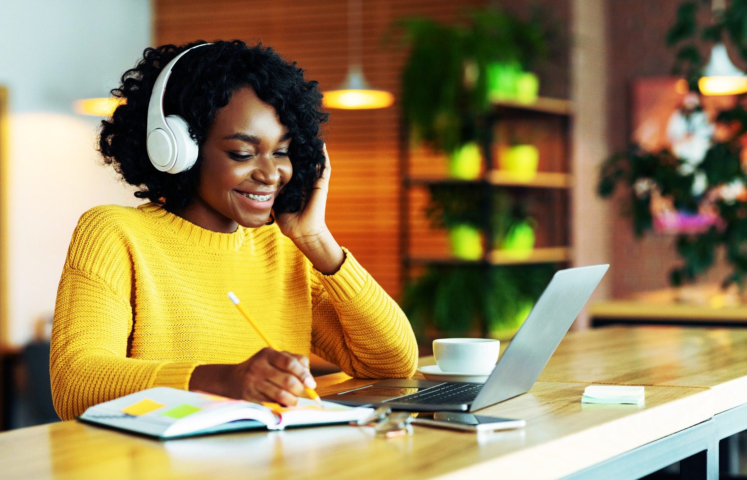 Black woman wearing headphones looking at laptop, smiling, and writing something down.
